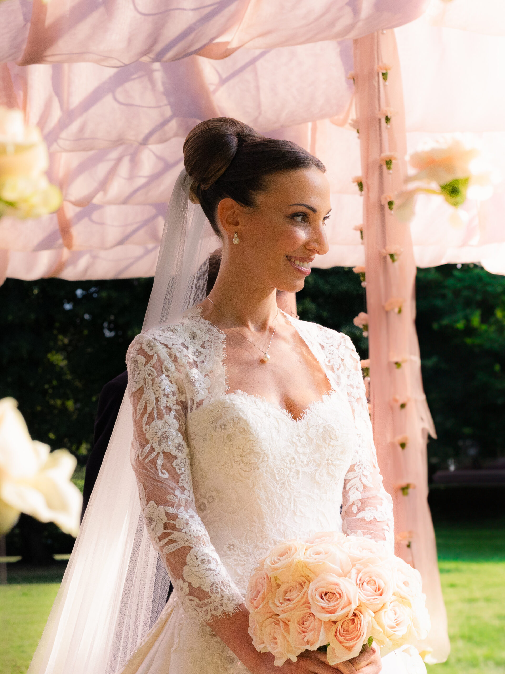 A beautiful bride in a lace wedding gown with long sleeves, holding a bouquet of pale pink roses, smiles radiantly under a pink canopy during her wedding in Milan. The bride's hair is styled in an elegant updo, and she wears a delicate veil that cascades down her back. This stunning moment is captured by Jan Schick Photography, a wedding photographer based in Switzerland.