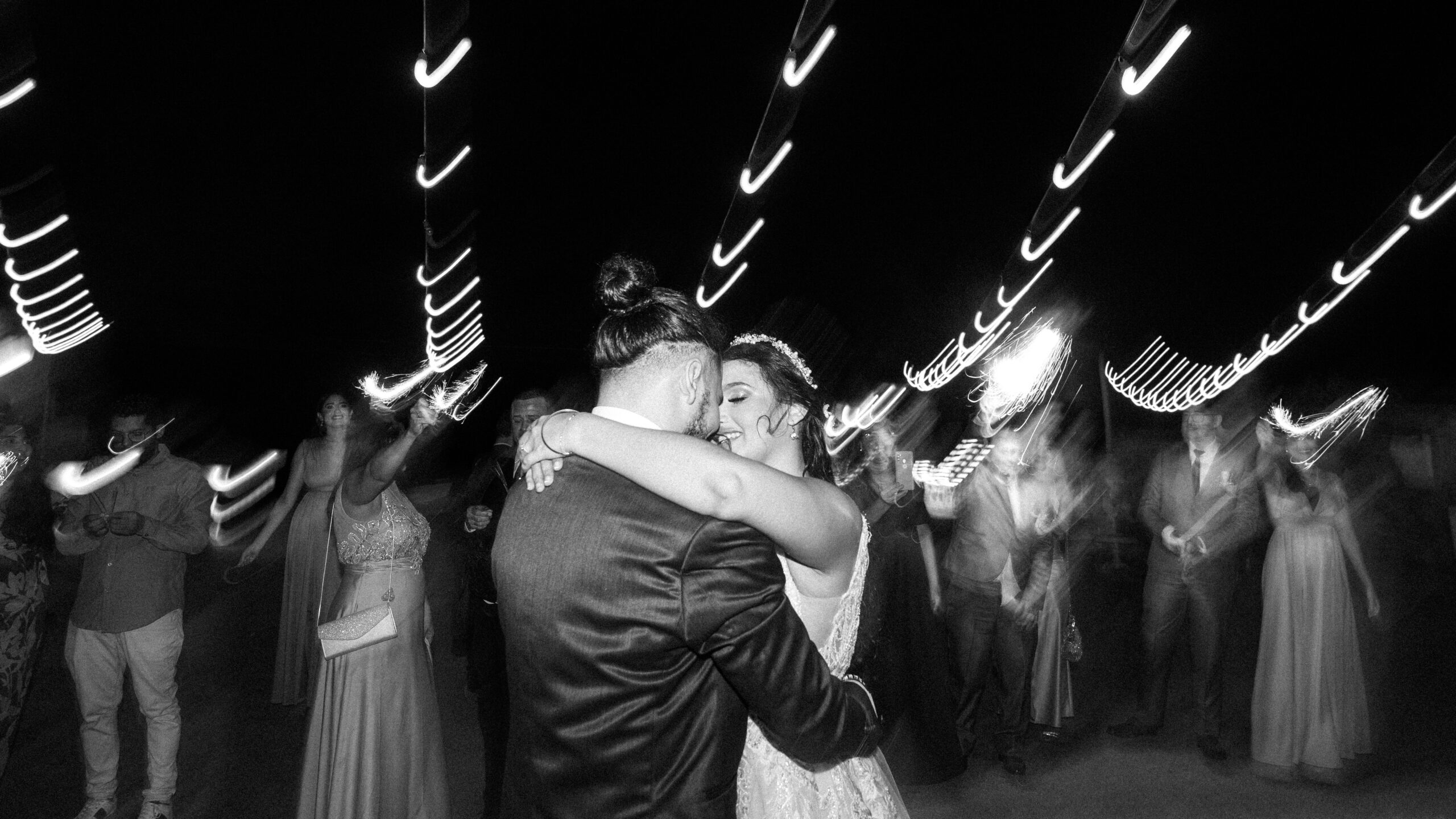 The newlyweds dance closely embraced under twinkling lights, surrounded by their guests. The movements and lights create an enchanting atmosphere that captures the joy and romance of this special moment. Das Brautpaar tanzt eng umschlungen unter funkelnden Lichtern, umgeben von ihren Gästen. Die Bewegungen und Lichter erzeugen eine bezaubernde Atmosphäre, die die Freude und Romantik dieses besonderen Augenblicks einfängt.