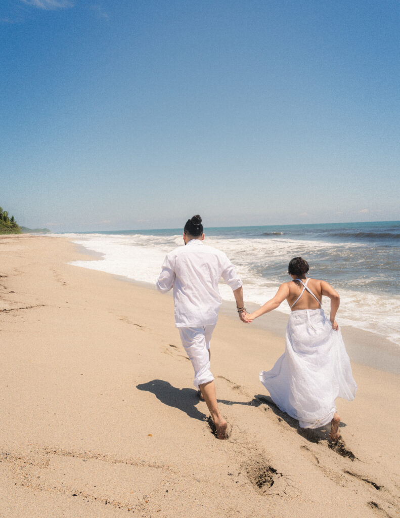 The bride and groom run hand-in-hand along the sandy shores of Tayrona National Park, radiating joy and connection during their elopement. Braut und Bräutigam laufen Hand in Hand entlang der sandigen Küste des Tayrona-Nationalparks und strahlen Freude und Verbundenheit während ihres Elopements aus.