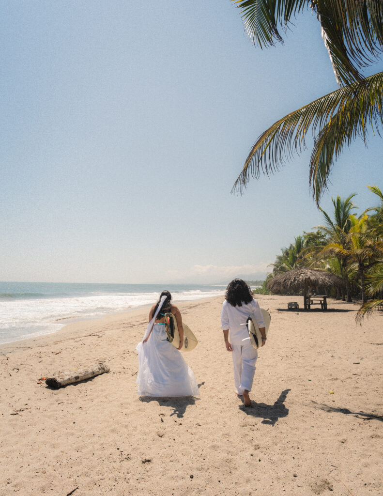 A couple dressed in white strolls along the pristine beach of Parque Tayrona, Colombia, carrying surfboards and embracing the tropical atmosphere. Captured by Jan Schick Photography. Ein Paar in Weiß spaziert entlang des unberührten Strandes im Parque Tayrona, Kolumbien, mit Surfbrettern und genießt die tropische Atmosphäre. Festgehalten von Jan Schick Photography.