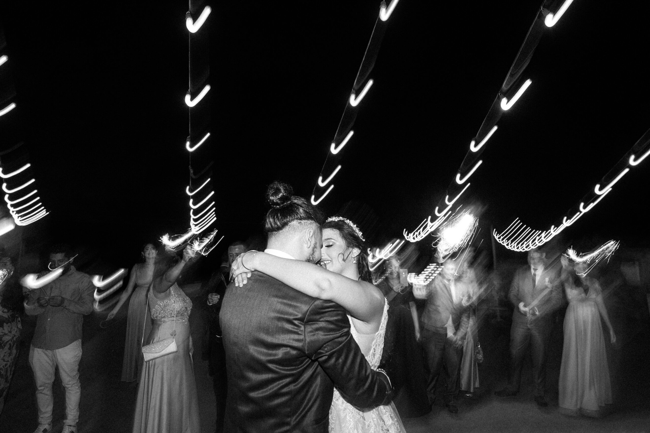 The newlyweds dance closely embraced under twinkling lights, surrounded by their guests. The movements and lights create an enchanting atmosphere that captures the joy and romance of this special moment. Das Brautpaar tanzt eng umschlungen unter funkelnden Lichtern, umgeben von ihren Gästen. Die Bewegungen und Lichter erzeugen eine bezaubernde Atmosphäre, die die Freude und Romantik dieses besonderen Augenblicks einfängt.