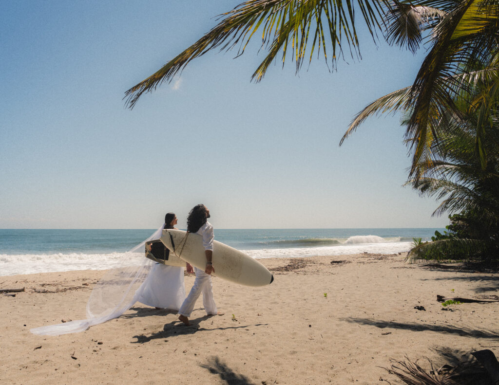 The couple walks along a sandy beach under swaying palm trees, their surfboards in hand, showcasing a unique elopement style in Parque Tayrona. Photography by Jan Schick. – Das Paar spaziert unter schwingenden Palmen mit ihren Surfbrettern in der Hand entlang eines Sandstrandes und zeigt einen einzigartigen Elopement-Stil im Parque Tayrona. Fotografie von Jan Schick.