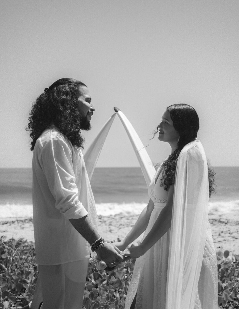 A tender moment as the couple exchanges vows by the ocean, standing under a symbolic surfboard arch in Parque Tayrona. Photography by Jan Schick. – Ein zärtlicher Moment, als das Paar am Ozean seine Gelübde unter einem symbolischen Surfbrettbogen im Parque Tayrona austauscht. Fotografie von Jan Schick.