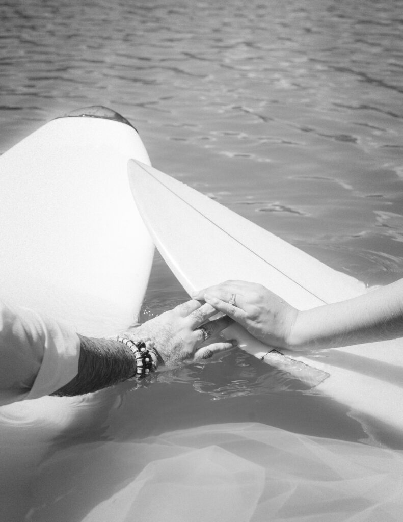 A close-up of the couple’s hands holding wedding rings and surfboards, symbolizing their adventurous love story in Parque Tayrona, Colombia. Black-and-white photography by Jan Schick. – Eine Nahaufnahme der Hände des Paares mit Eheringen und Surfbrettern, die ihre abenteuerliche Liebesgeschichte im Parque Tayrona, Kolumbien, symbolisieren. Schwarz-Weiß-Fotografie von Jan Schick.